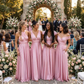 color:Blushing Pink |  Four bridesmaids in matching floor-length blush pink off-the-shoulder and V-neck dresses standing together outdoors at a wedding ceremony, with guests, white chairs, and floral arch decorations in the background.